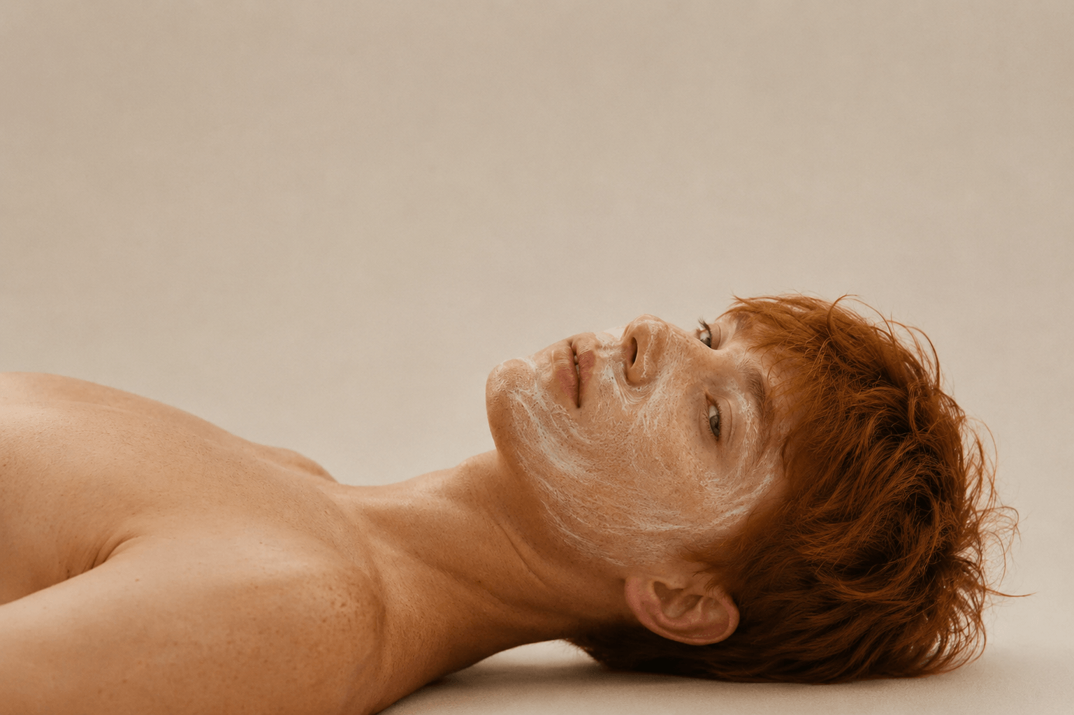 Male skincare model with red hair and freckles relaxing with a facial mask treatment, photographed against a neutral background to showcase self-care, hydration and healthy-looking skin