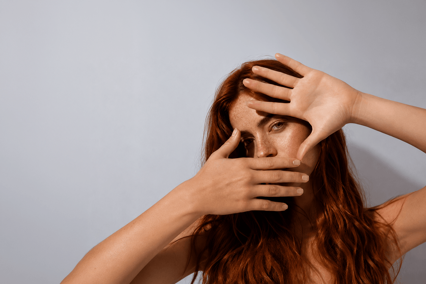 Freckled red-haired woman framing her face with her hands, highlighting natural skin texture and a quiet, minimalist beauty aesthetic