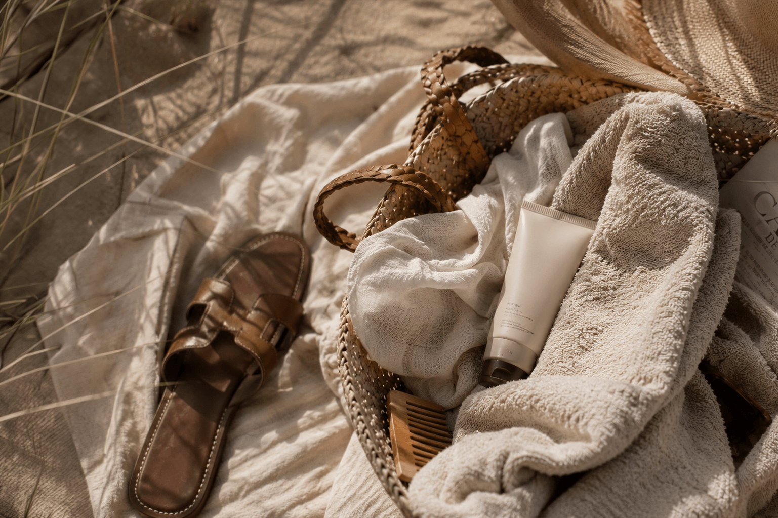 Thoughtfully arranged summer essentials on a sandy beach, featuring a woven straw tote, soft linen textiles, a minimalist SPF skincare product, leather sandals and a folded newspaper, captured in warm natural light with a refined coastal atmosphere