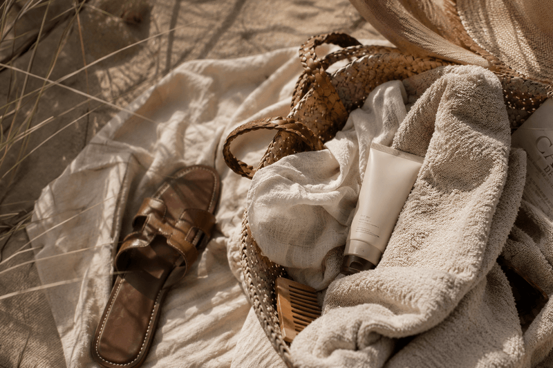 Thoughtfully arranged summer essentials on a sandy beach, featuring a woven straw tote, soft linen textiles, a minimalist SPF skincare product, leather sandals and a folded newspaper, captured in warm natural light with a refined coastal atmosphere