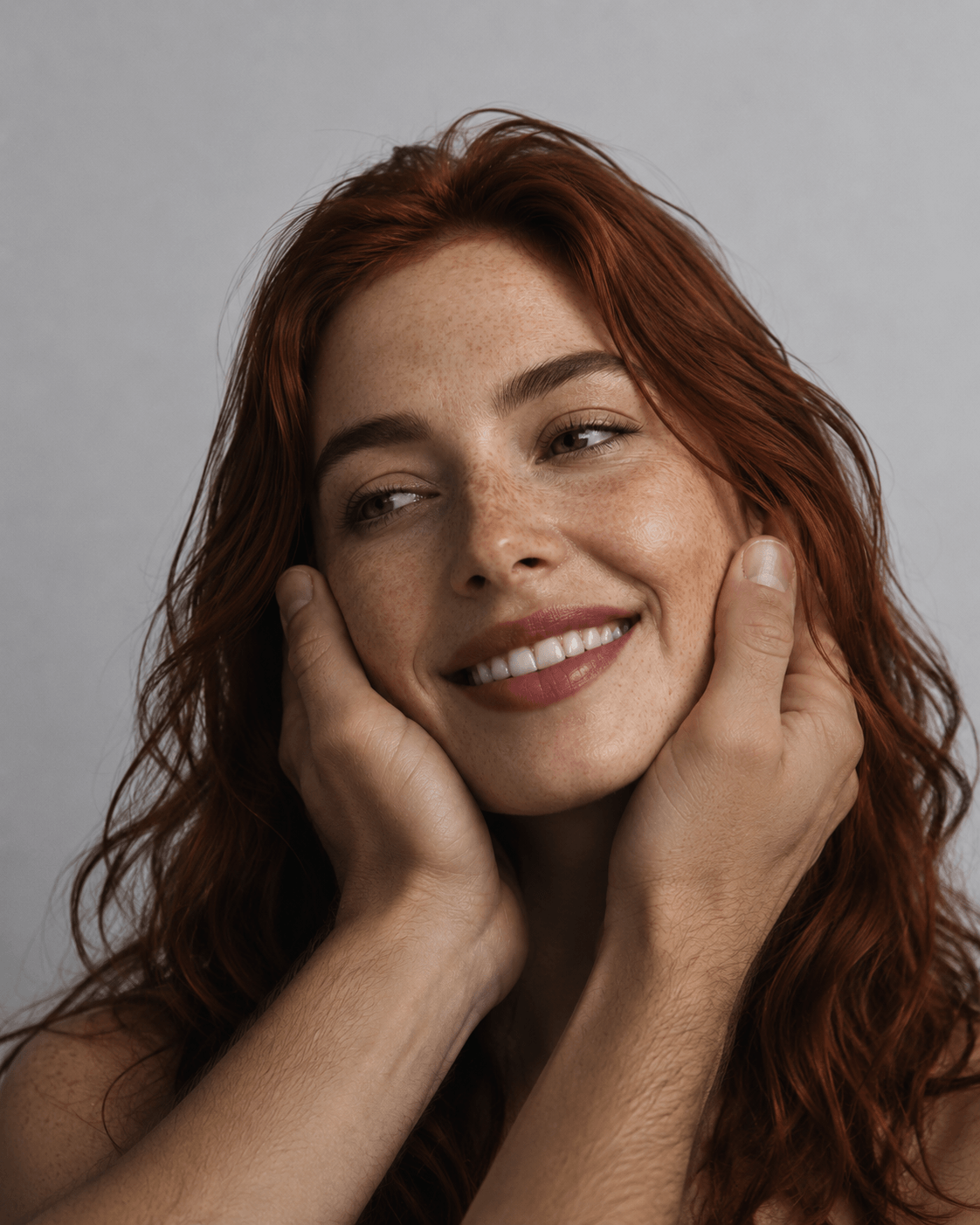 Woman with red hair smiling and touching her face against a gray background - vegan skincare ritual by Suvera Skincare