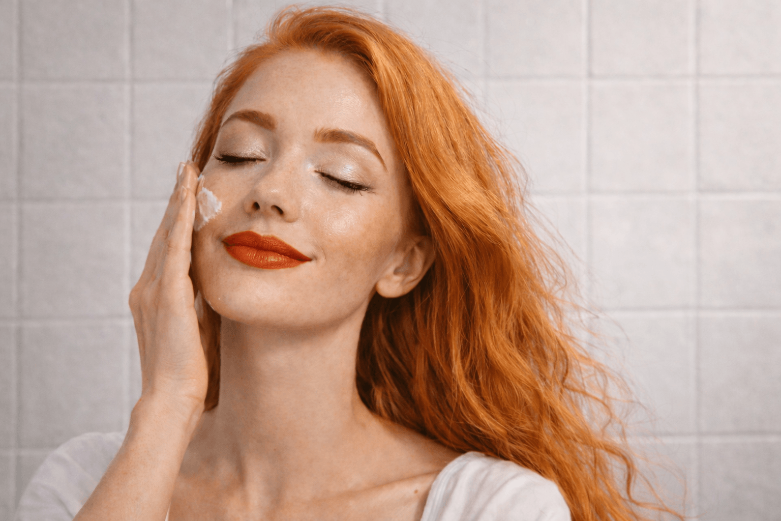 Woman with red hair applying cream to her face against a tiled wall background
